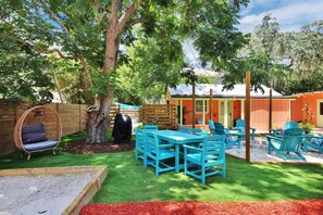 Ein Garten mit einer Holzterrasse, einem blauen Tisch und Stühlen, einem Schaukelstuhl und einem Grill.