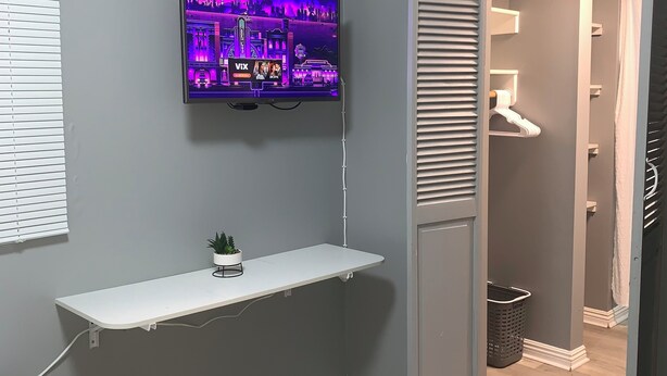 Een moderne badkamer met een grijze wand, een tv aan de muur met een stadsgezicht, een witte plank met een plant, en een deur naar een douche.