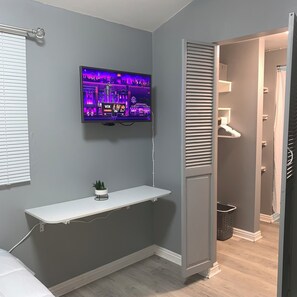 A modern bathroom with a grey wall, a mounted TV displaying a cityscape, a white shelf with a plant, and a door leading to a shower area. - The Small Sandalwood -"Sandalito"· Private Suite, curated for comfort & privacy. (El Centro)