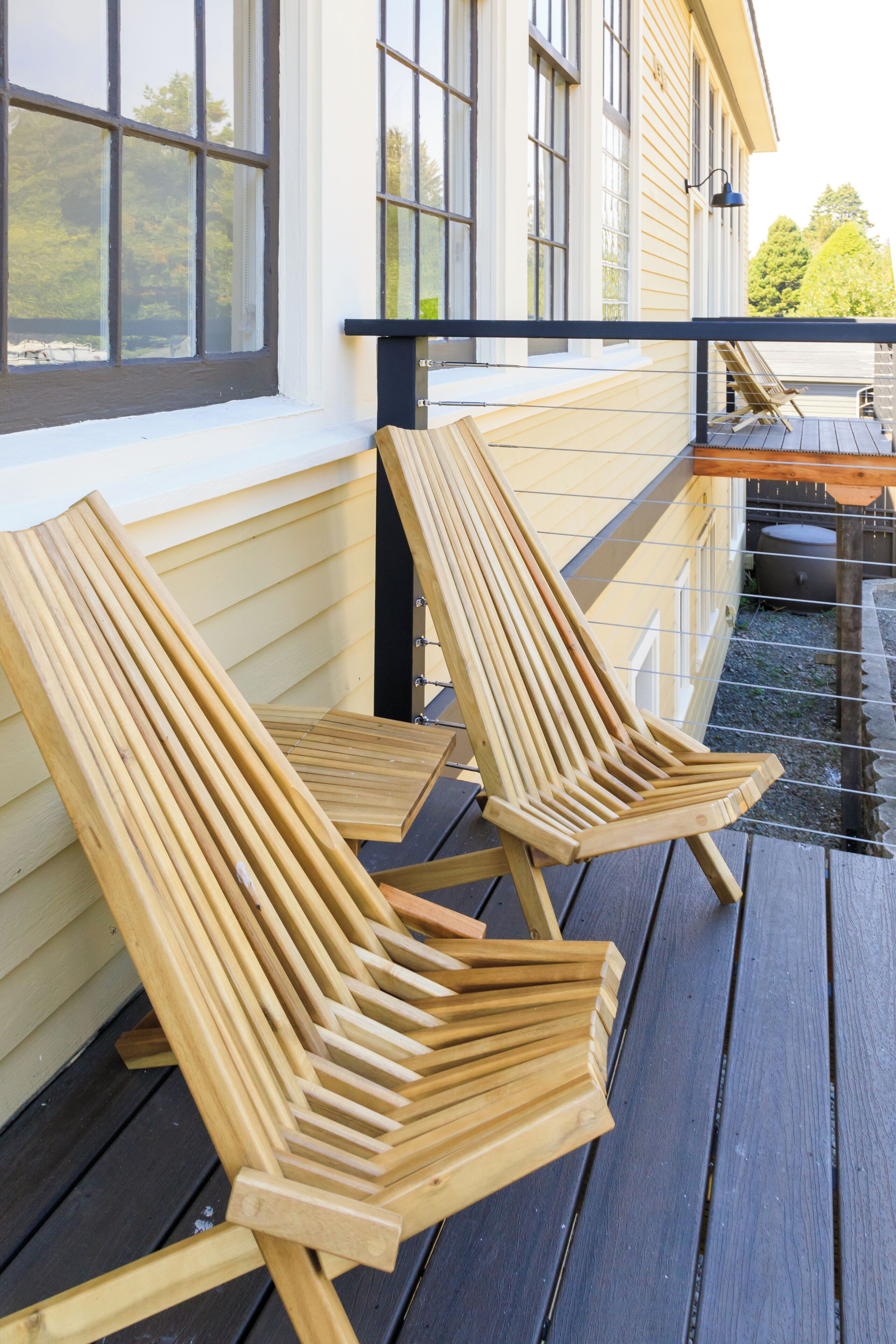 Two wooden Adirondack chairs on a wooden deck with a railing, next to a building with large windows.