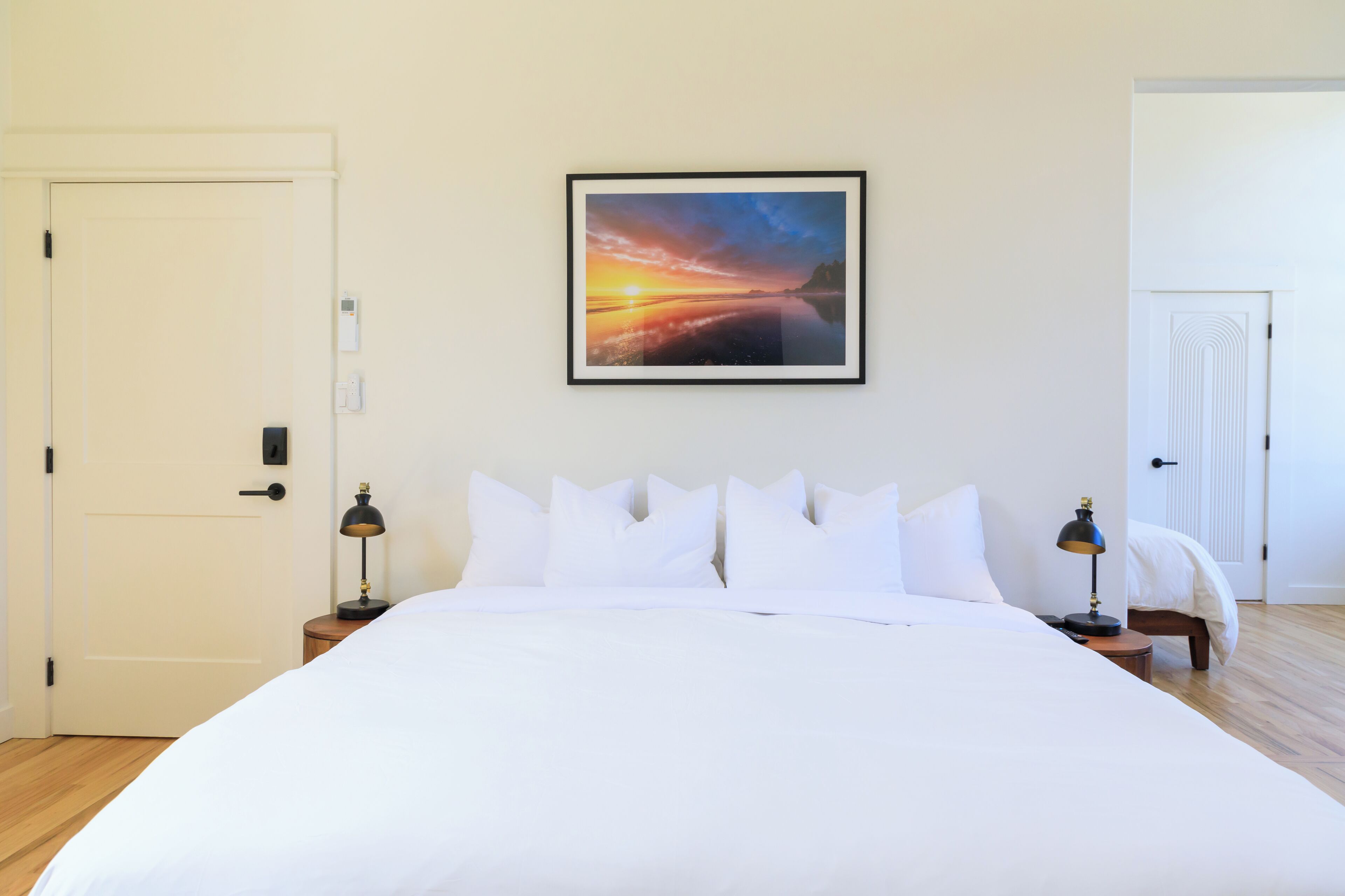 A neatly made bed with white linens, a framed sunset painting on the wall, and two bedside lamps.