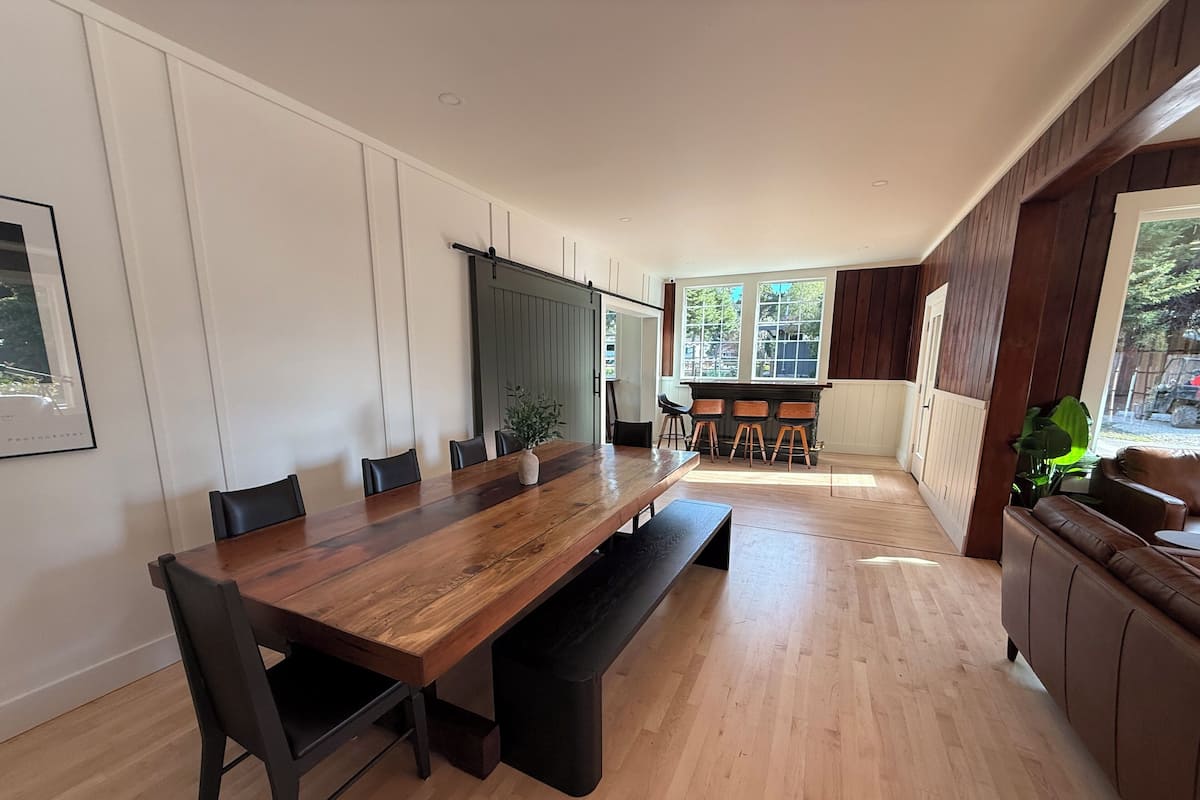 A modern dining area with a long wooden table, black chairs, and a bench. There are three wooden stools at a small table near the window. The room has white walls, wooden paneling on the upper part, and a view of greenery outside.
