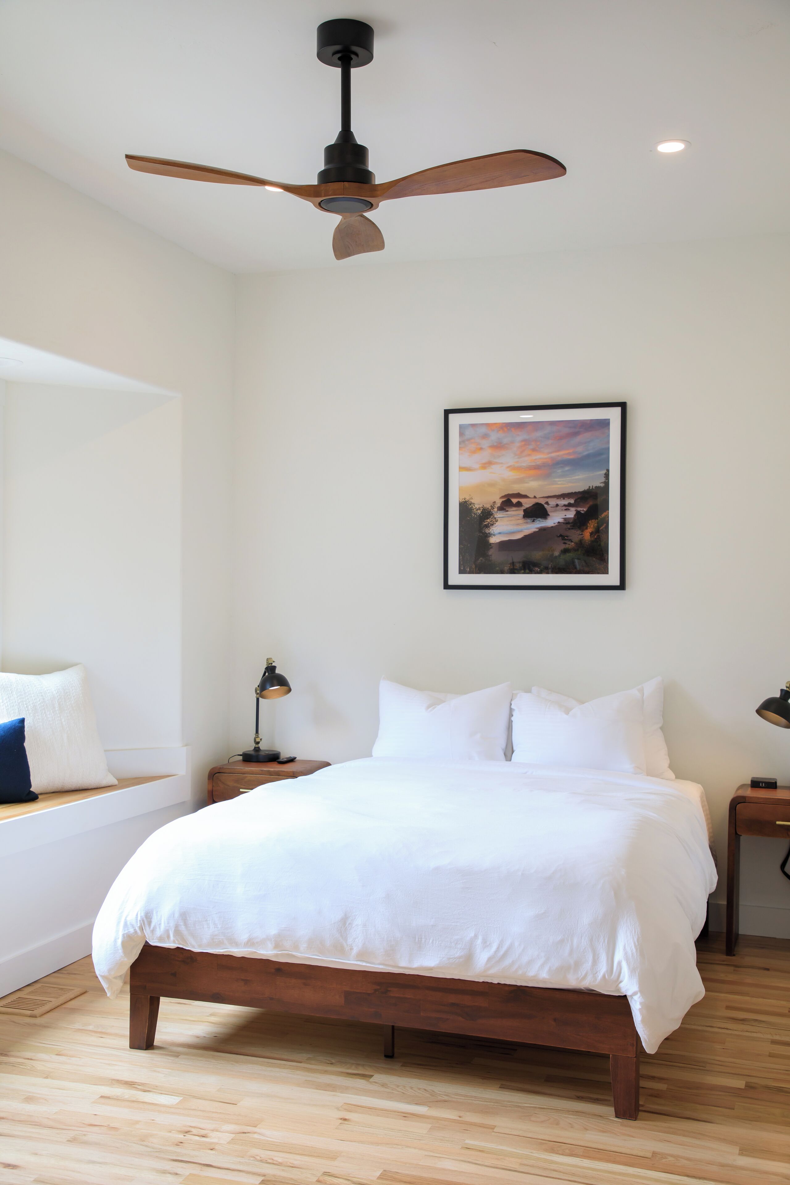Un dormitorio con una cama de madera, un ventilador de techo, una mesita de noche con una lámpara y un cuadro en la pared.