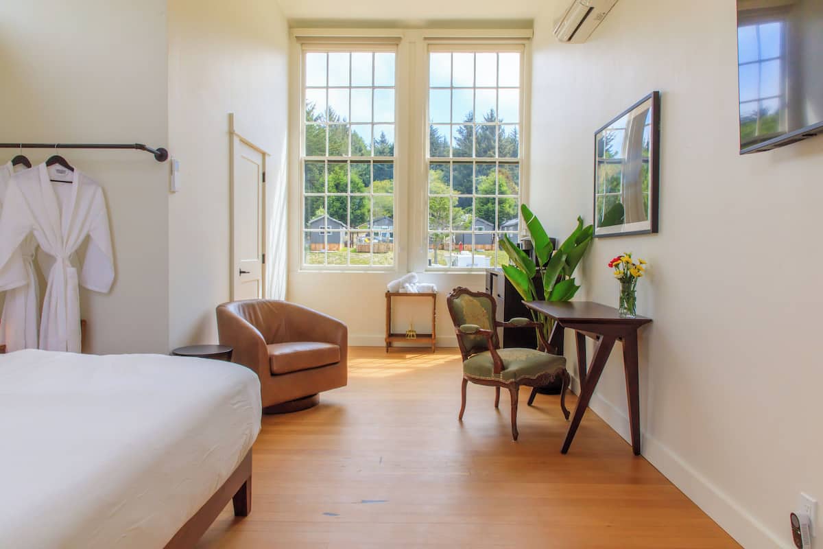A hotel room with a bed, a chair, a table with a vase, and a window with a view of houses and trees.