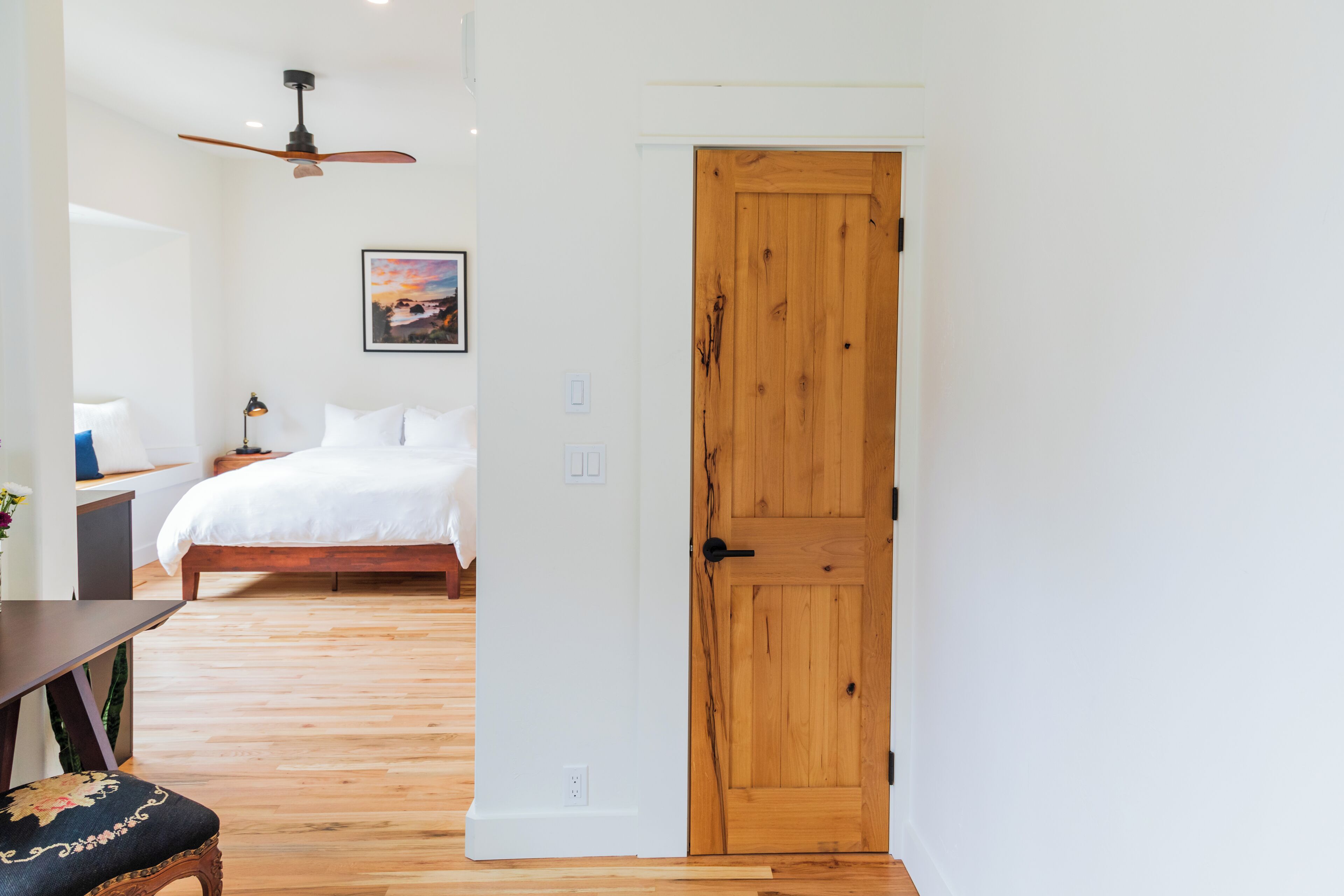 A bedroom with a wooden door, a bed, a ceiling fan, and a bedside table with a lamp.