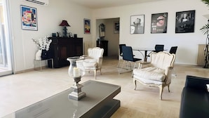 En moderne stue med et glasbord, hvide lænestole og en mørk sofa. Væggene er pyntet med billeder i rammer, der er et mørkt træskab, og et spiseområde med bord og stole.