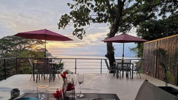 An outdoor dining area with tables and chairs, two umbrellas, and a view of the sea. - Suite 4 Hotel VOS - Superior Suite, Ocean View (Manuel Antonio)
