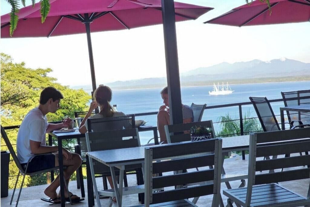 Un área de comedor al aire libre con mesas y sillas, sombrillas y vistas al mar y montañas de fondo.