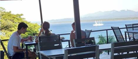 Un área de comedor al aire libre con mesas y sillas, sombrillas y vistas al mar y montañas de fondo.