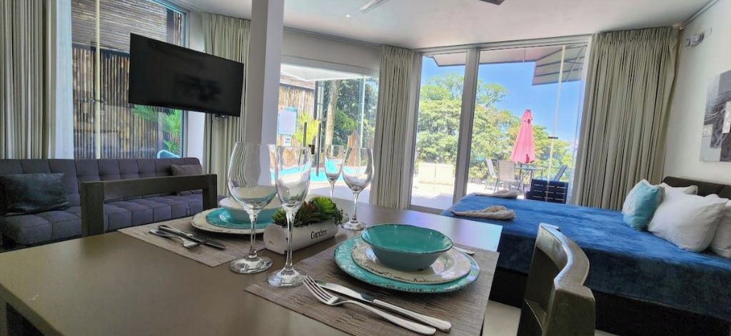 A modern hotel room with a dining area, a sofa, a TV mounted on the wall, and a balcony with a view of the outdoors.