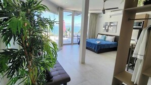 A modern hotel room with a balcony, a bed, a seating area, and a large plant.
