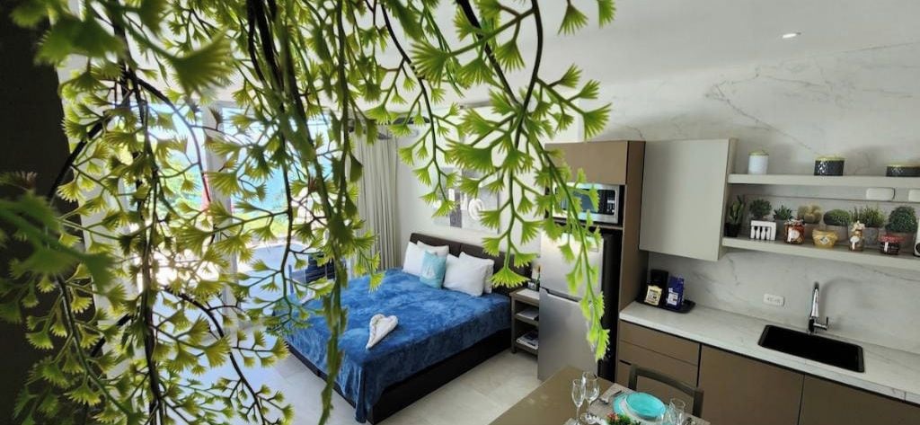 A modern hotel room with a bed, a desk, a kitchen area, and a view of the outdoors.