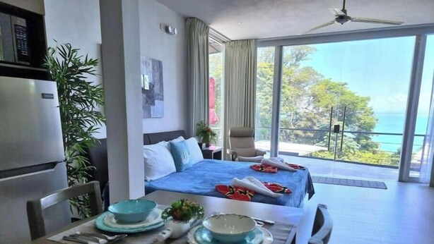 Une chambre d’hôtel avec un lit, une table à manger avec des assiettes et des bols, une chaise, un ventilateur de plafond et une vue sur l’extérieur.
