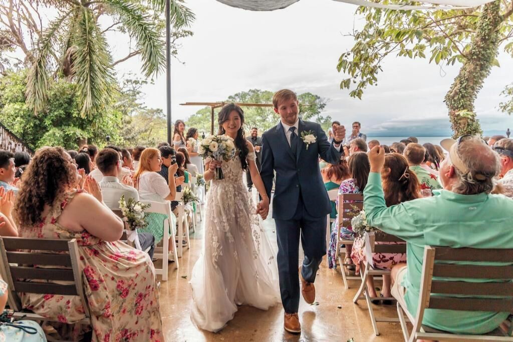A wedding ceremony with a couple walking down the aisle, surrounded by guests seated on chairs.