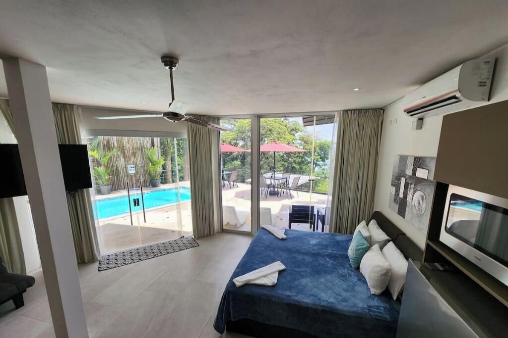 Ein modernes Hotelzimmer mit einem großen Bett, einem Flachbildfernseher und Blick auf einen Swimmingpool sowie eine Außenbereich-Sitzgelegenheit.