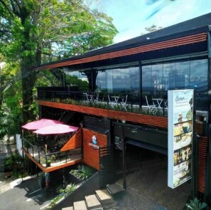 Un bâtiment moderne avec une façade en verre, un espace extérieur aménagé pour les clients et une enseigne publicitaire pour un restaurant.