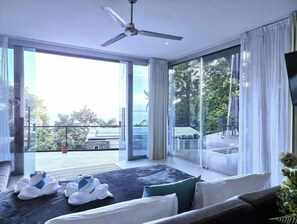 Une chambre d’hôtel moderne dotée d’une grande porte-fenêtre coulissante donnant sur un balcon, d’un ventilateur de plafond et d’un lit agrémenté de serviettes blanches et bleues.