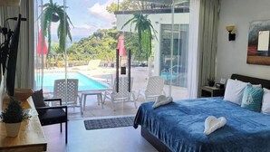 A hotel room with a large bed, a desk, and a view of a pool and outdoor seating area. - Suite 7 Hotel VOS - Superior Suite, Ocean View (Manuel Antonio)