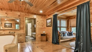 A wooden cabin interior with a bed, a bathroom, and a large window with curtains.
