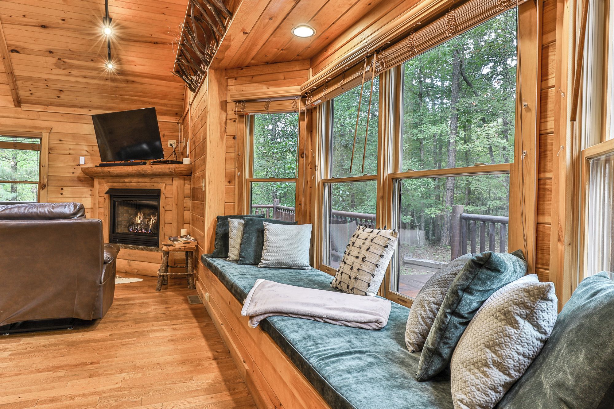 A log cabin interior with a fireplace, a sofa, and large windows offering a view of the forest.