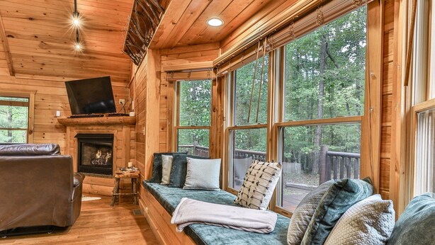 A cozy cabin interior with a fireplace, a large window overlooking a forest, and a comfortable seating area.