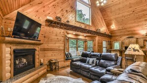 A cozy log cabin interior with a fireplace, leather sofa, and a television mounted on the wall.