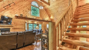 A log cabin interior with a fireplace, leather sofa, wooden staircase, and large windows.