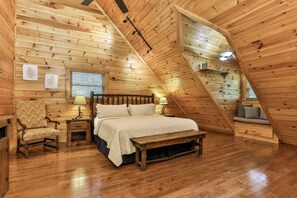 Een knus zolderkamer met een houten schuine plafond, een bed met een witte spreideken, een bankje, een stoel en een kleine tafel met een lamp.