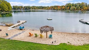 Une plage au bord d’un lac, agrémentée d’un parasol en chaume, de chaises Adirondack aux couleurs vives et d’un quai où sont amarrés des bateaux.