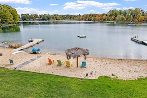 A lakeside beach with a thatched umbrella, colorful Adirondack chairs, and a dock with boats.