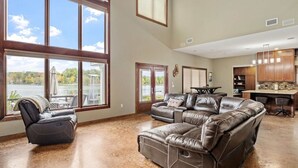 Un salon spacieux, doté d’un grand canapé d’angle en cuir, d’un fauteuil inclinable et offrant une vue sur un lac grâce à de grandes fenêtres.