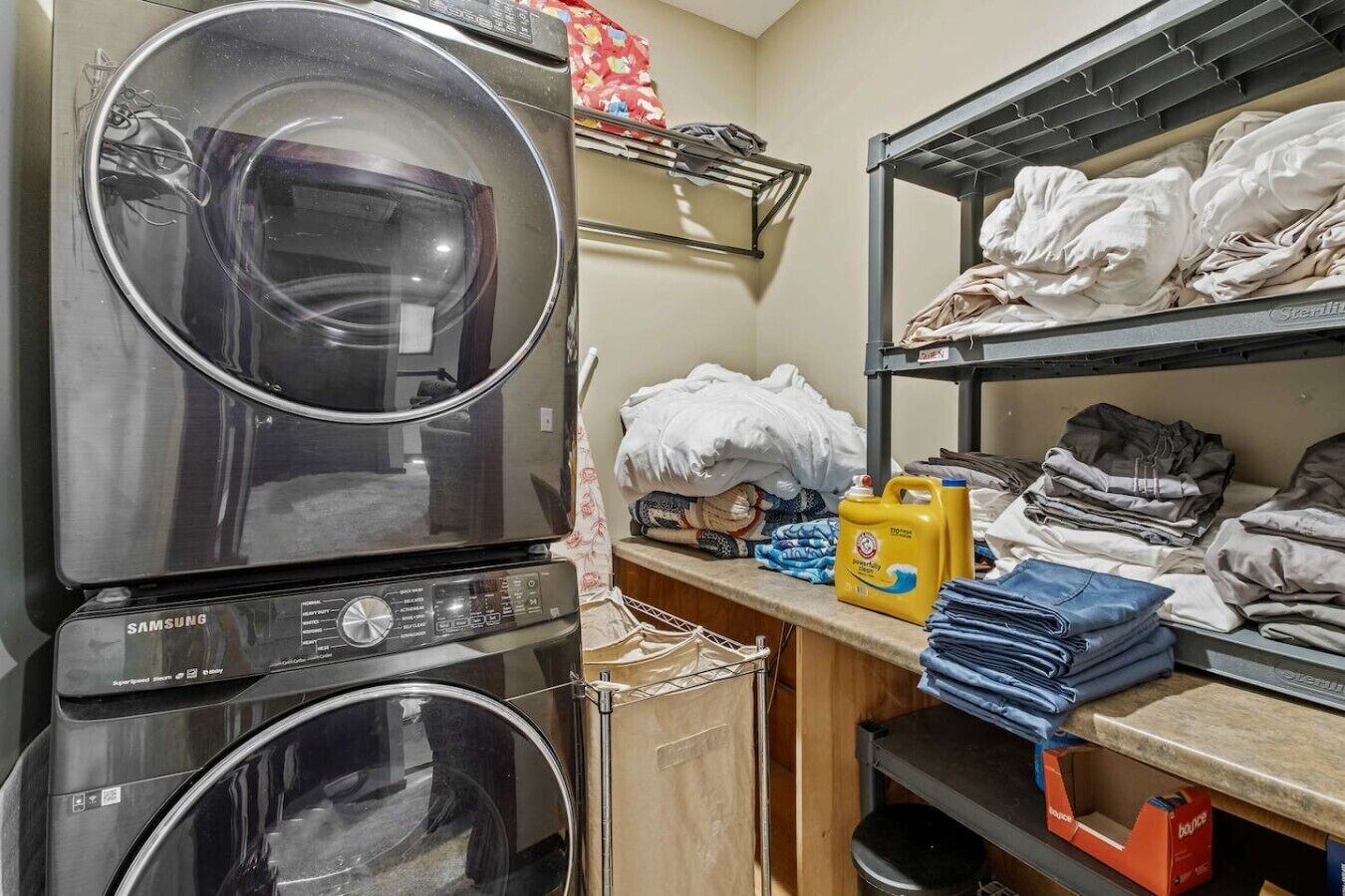 Uma área de lavandaria com máquinas de lavar e secar empilhadas, prateleiras com roupas dobradas e uma garrafa de detergente.