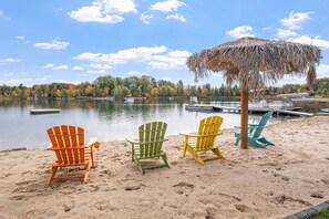 A beach with colorful Adirondack chairs, a thatched umbrella, and a calm lake with boats.