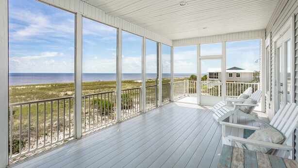 A covered porch with a view of the ocean, white railings, and two white chairs with cushions.