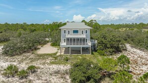A two-story house with a balcony and a covered porch, surrounded by dense vegetation and sandy terrain.
