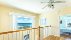 A room with a wooden floor, a ceiling fan, and a view of the ocean through large windows.