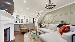 A spacious living area with a white sofa, a patterned armchair, and a glass coffee table. A staircase leads to an upper level, and a chandelier hangs from the ceiling.