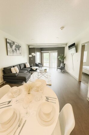 A modern living room with a dining area set for a meal, a black sofa, a white dining table, and a large window.
