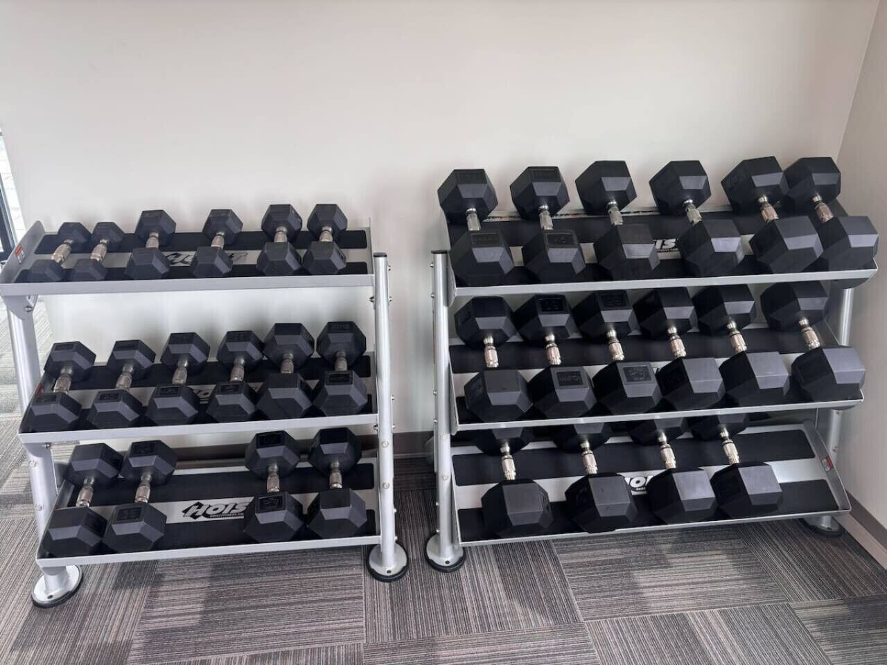 A rack of hexagonal dumbbells with weight markings, arranged on a metal stand with wheels, against a plain wall.
