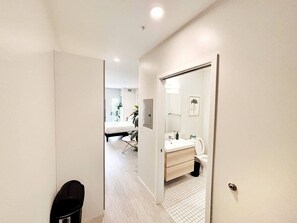 Un couloir mène à une salle de bain équipée de toilettes, d’un lavabo et d’un miroir. On aperçoit un lit au fond.