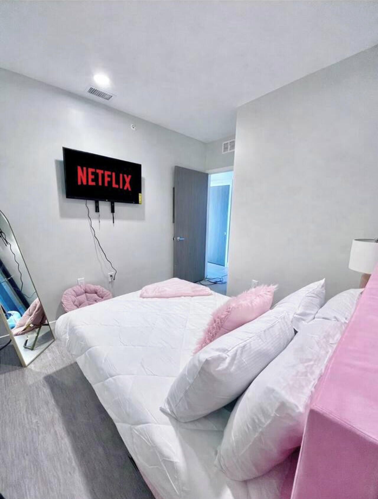 A modern bedroom with a large bed, a wall-mounted TV displaying 'NETFLIX,' and a door leading to another room.