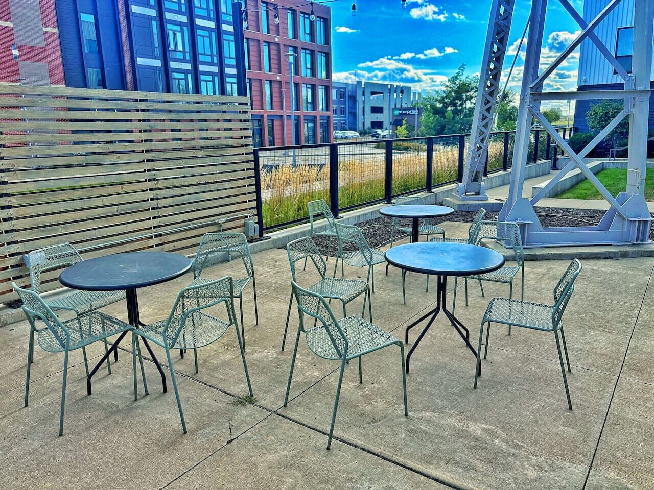 An outdoor patio with metal tables and chairs, a wooden fence, and a view of buildings in the background.