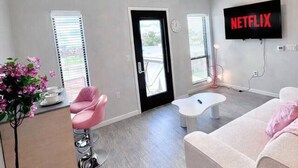 Ein modernes Wohnzimmer mit einer Couch, einem Couchtisch, einem rosa Sessel, einem an der Wand montierten Fernseher und einer Tür, die nach draußen führt.