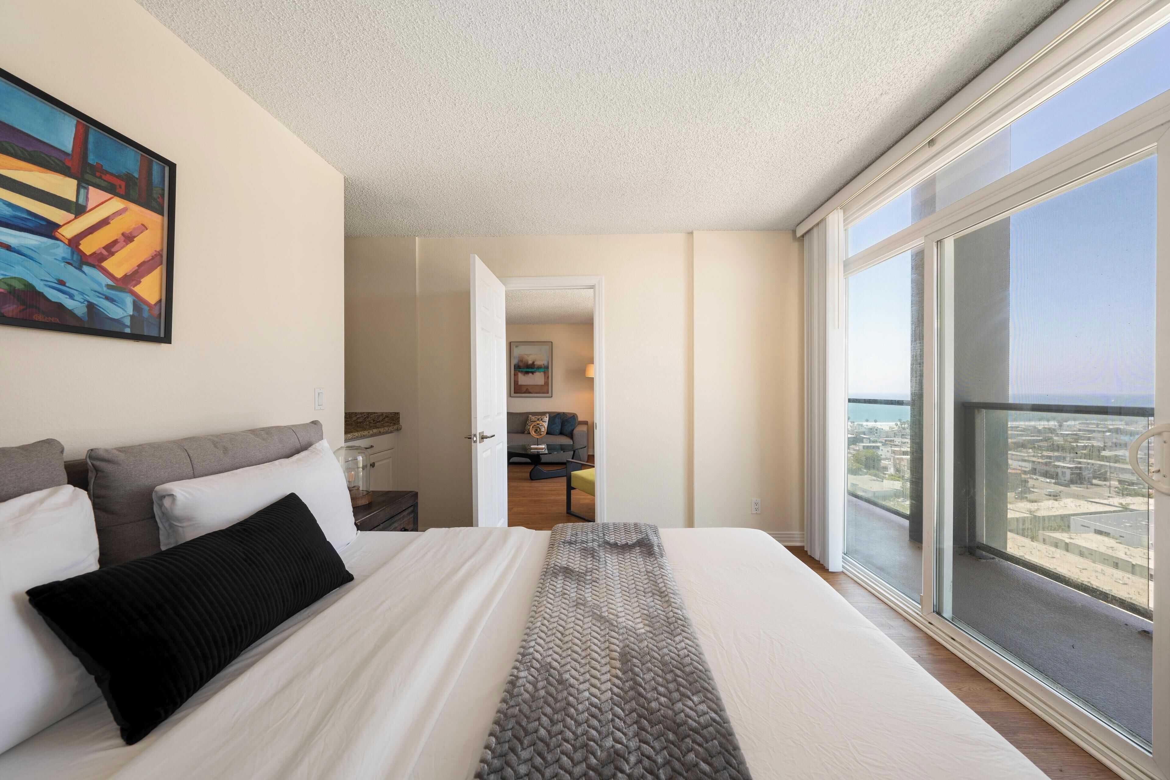 A bedroom with a large bed, a view of the city through the window, and a painting on the wall.