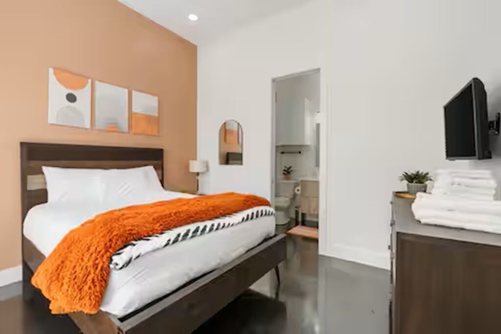 Une chambre d’hôtel moderne dotée d’un grand lit, d’un téléviseur à écran plat fixé au mur et d’une salle de bain visible à travers une porte ouverte.