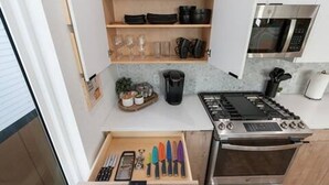 Une cuisine compacte équipée d’une cuisinière, d’un four et d’un four à micro-ondes intégrés. On y trouve un tiroir contenant divers ustensiles et une étagère avec des assiettes et des tasses.