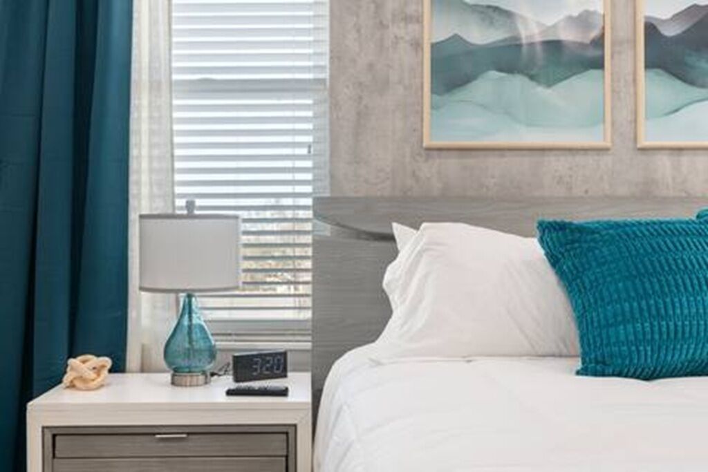 A bedroom with a bed, a nightstand with a lamp, a clock, and artwork on the wall.
