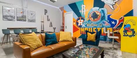 A room with a brown leather sofa, a coffee table with board games, a colorful mural with a baseball theme, and a dining area with a white table and blue chairs.