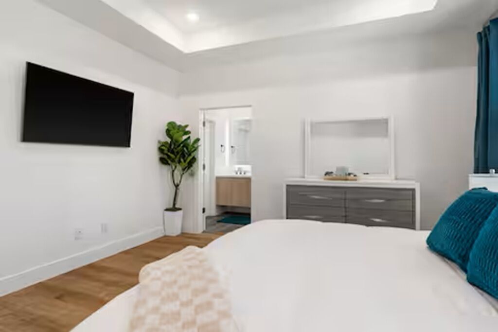 A modern bedroom with a large bed, a flat-screen TV mounted on the wall, a dresser, and a potted plant.