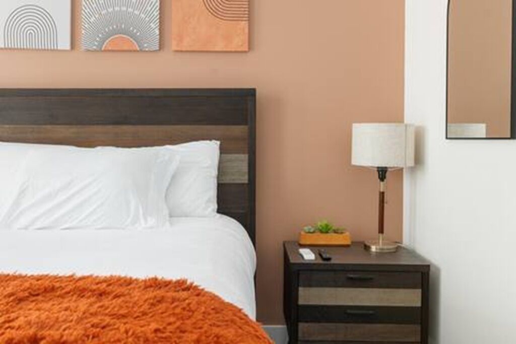 Un lit bien fait, avec du linge de lit blanc et un plaid orange, une lampe de chevet et une petite table sur laquelle se trouvent une plante et une télécommande.